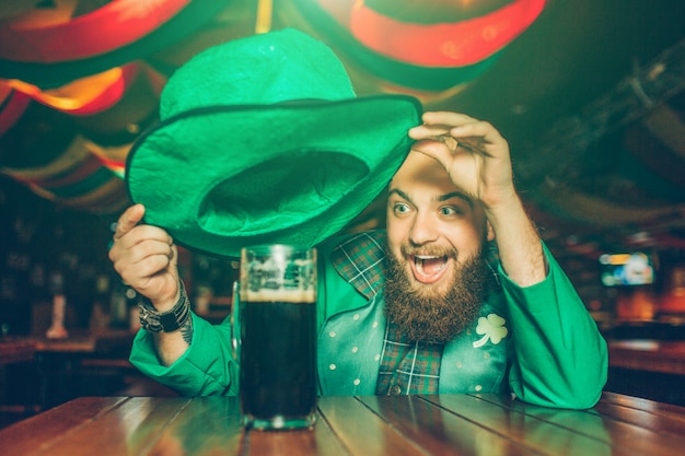 Il giovane emozionante felice in vestito verde si siede alla tavola in pub. Tiene il cappello sopra un boccale di birra scura. Sorriso del giovane