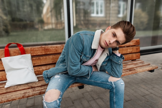 Il giovane europeo in giacca di jeans alla moda in maglietta rosa in jeans vintage con shopper in tessuto si siede su una panca di legno alla fermata dell'edificio pubblico in città. Ragazzo attraente in abiti alla moda per strada.