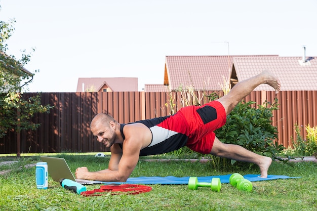 Il giovane fa sport all'aperto. Lo sportivo allegro con i capelli neri fa gli affondi sul prato in giardino, accanto c'è un laptop con allenamento online