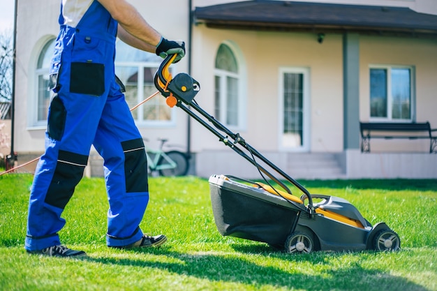 Il giovane falcia il prato usando un tosaerba elettrico in una tuta da lavoro speciale vicino a una grande casa di campagna nel cortile sul retro