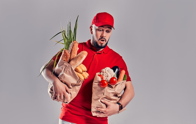 Il giovane fattorino barbuto in uniforme rossa tiene due grandi pacchi pesanti con pane e verdure isolati su sfondo grigio.