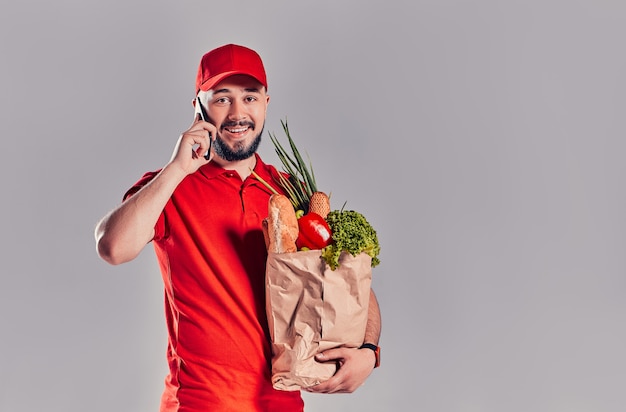 Il giovane fattorino barbuto in uniforme rossa tiene il pacchetto con pane e verdure e parla su smartphone isolato su sfondo grigio.