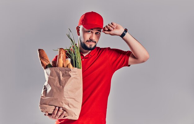 Il giovane fattorino barbuto in uniforme rossa tiene il pacchetto con pane e verdure isolati su sfondo grigio.