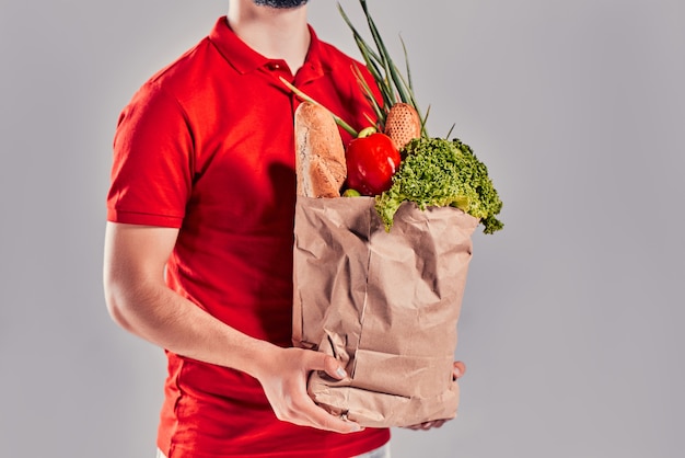 Il giovane fattorino barbuto in uniforme rossa tiene il pacchetto con pane e verdure isolati su sfondo grigio.