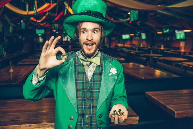 Il giovane felice nel vestito verde sta in pub e tiene le monete dorate. Ne guarda uno e stupisce. Il ragazzo indossa l'abito di San Patrizio.