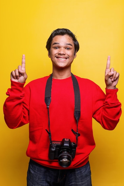 Il giovane fotografo eccitato porta la fotocamera e punta il dito verso l'alto isolato su sfondo di colore giallo