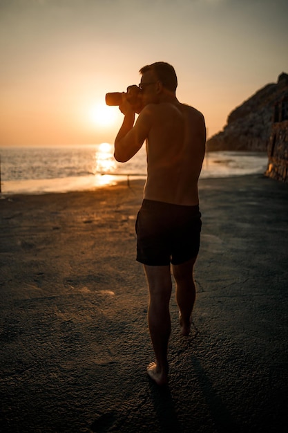Il giovane fotografo maschio fa fotografie del mare al tramonto stando in piedi sulla riva. Tramonto sul mare. Messa a fuoco selettiva. Il turista fotografa il paesaggio marino