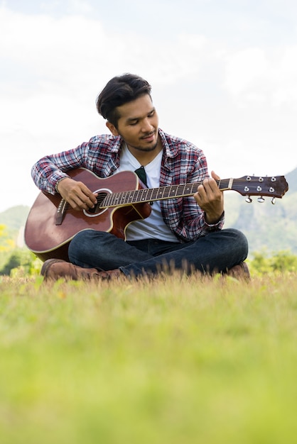 Il giovane gioca la chitarra mentre si siede sull&#39;erba verde contro la natura e il paesaggio della montagna