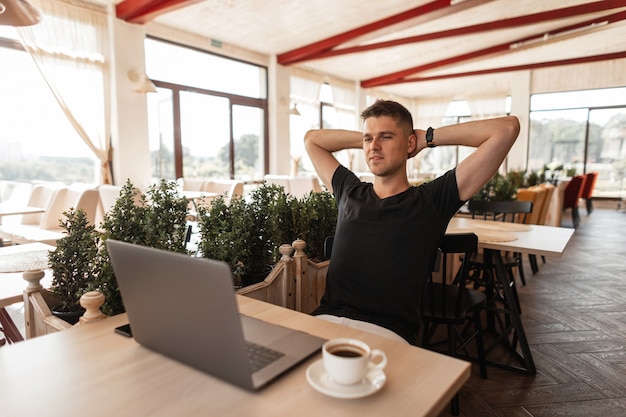 Il giovane gioioso di successo in una maglietta nera con un computer si siede in un caffè moderno. Ragazzo libero professionista felice che lavora a distanza su un computer portatile. Tempo di relax.