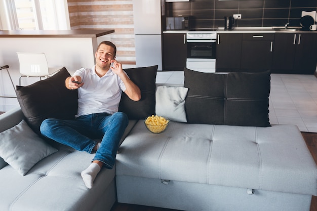 Il giovane guarda la tv nel suo appartamento. Felice ragazzo eccitato parlando al telefono e utilizzando il telecomando per cambiare i canali TV