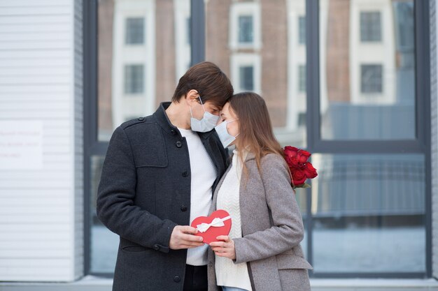 Il giovane ha sorpreso la sua ragazza con bouquet di rose e confezione regalo di cuore in città. Giorno di San Valentino. Stare in piedi e guardarsi l'un l'altro. Mascherina. Foto di alta qualità