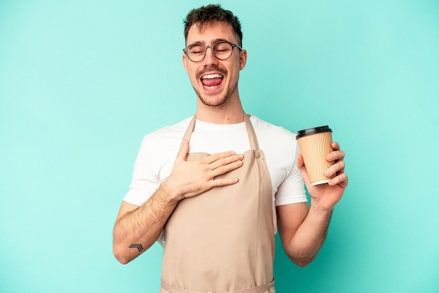 Il giovane impiegato di negozio che tiene un caffè isolato su sfondo blu ride ad alta voce tenendo la mano sul petto.