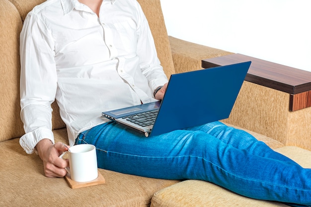 Il giovane in camicia bianca e blue jeans tiene la tazza di tè e lavora al computer portatile, scrive in taccuino in ufficio a casa seduto sul divano beige accogliente