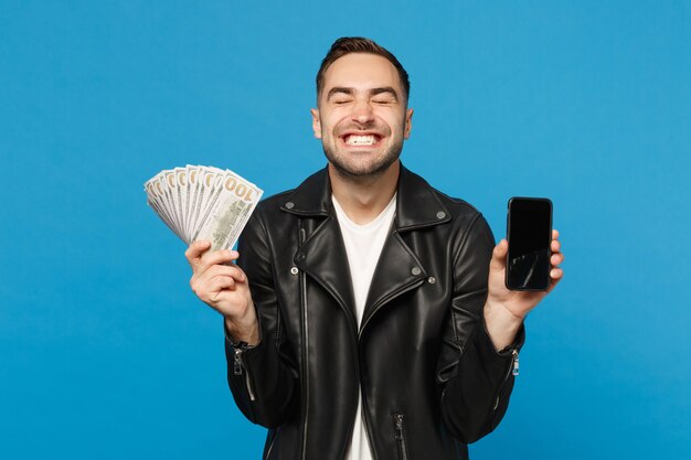 Il giovane in maglietta bianca della giacca nera tiene lo schermo vuoto del telefono cellulare per il denaro contante del contenuto promozionale isolato sul ritratto blu dello studio del fondo della parete. Concetto di stile di vita della gente. Mock up copia spazio