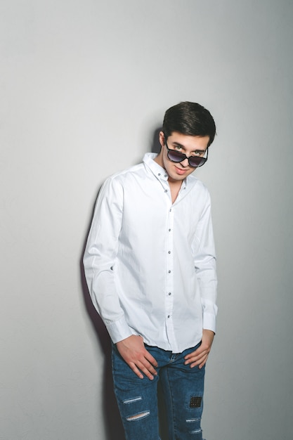 Il giovane in pantaloncini e maglietta bianca sta sorridendo in piedi vicino al muro con gli occhiali