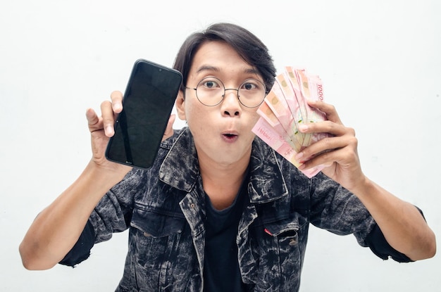 Il giovane indonesiano attraente felice ha scioccato felicemente mentre tiene la carta moneta e il telefono della rupia.