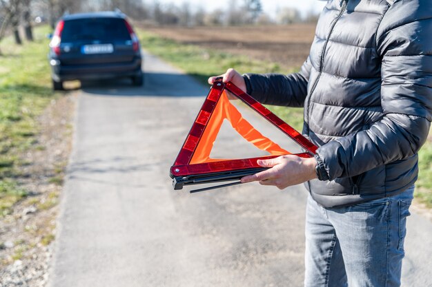 Il giovane installa un triangolo d'avvertimento arancione sulla strada davanti a un'auto rotta.