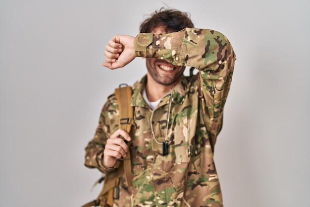 Il giovane ispanico che indossa l'uniforme dell'esercito mimetico sorridente allegro gioca a sbirciare un fischio con le mani che mostrano il viso sorpreso ed uscito