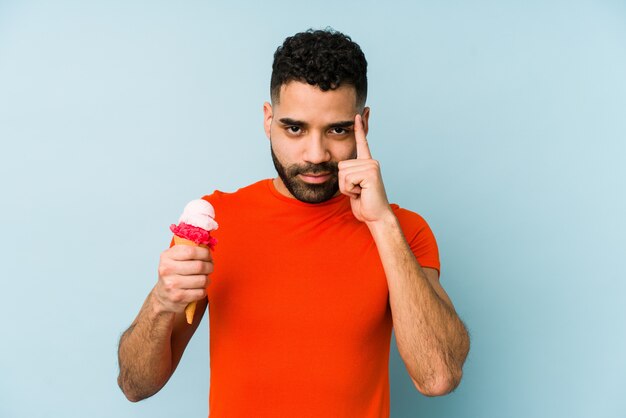 Il giovane latino che tiene un gelato che indica il tempio con il dito, pensando, si è concentrato su un compito.