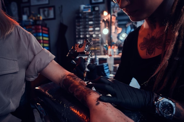 Il giovane maestro del tatuaggio concentrato sta facendo il tatuaggio sulla mano del cliente al salone del tatuaggio scuro.