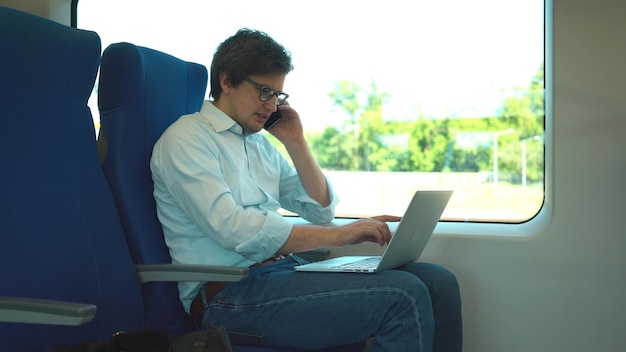 Il giovane manager che indossa una camicia bianca e jeans sta andando al lavoro in treno e sta usando il suo laptop