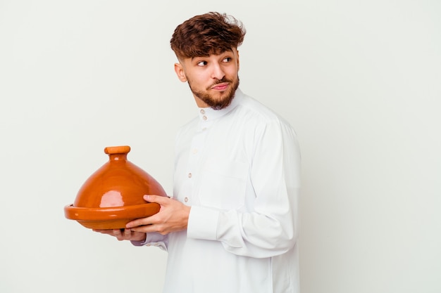 Il giovane marocchino che indossa il costume arabo tipico che tiene un tajine isolato su bianco guarda da parte sorridente, allegro e piacevole.
