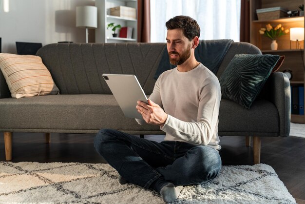 Il giovane maschio allegro è seduto in soggiorno nella posa del loto e guarda lo schermo del tablet nelle mani con interesse