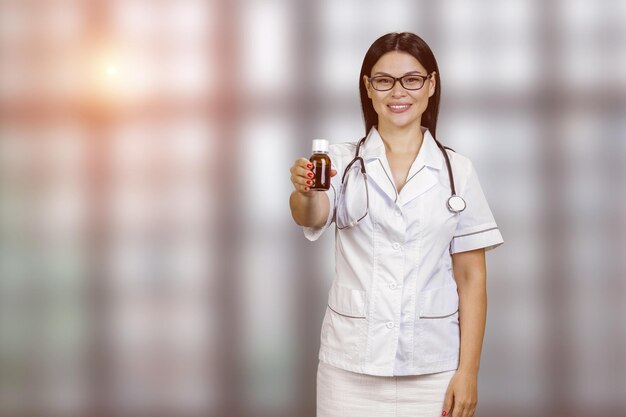 Il giovane medico asiatico femminile felice sta pubblicizzando la bottiglia della medicina ha offuscato il fondo a scacchi delle finestre