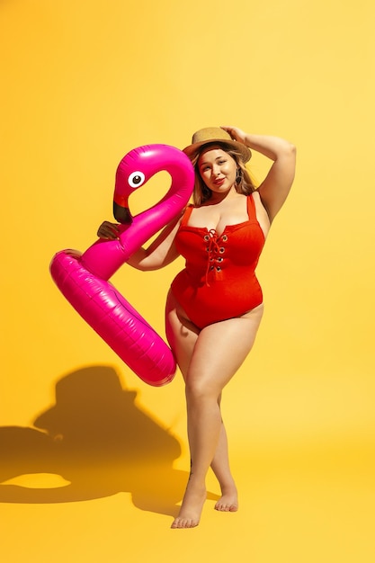 Il giovane modello femminile caucasico plus size si sta preparando per il resort sulla spiaggia su sfondo giallo. Donna in costume da bagno rosso e cappello in posa con swimring. Concetto di estate, festa, corpo positivo, uguaglianza.