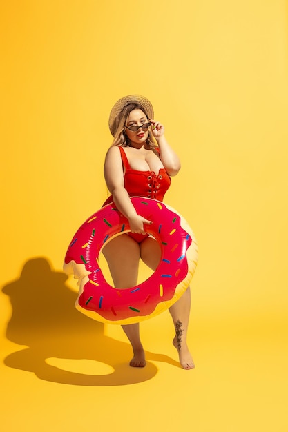 Il giovane modello femminile caucasico plus size si sta preparando per il resort sulla spiaggia su sfondo giallo. Donna in costume da bagno rosso e cappello in posa con swimring. Concetto di estate, festa, corpo positivo, uguaglianza.