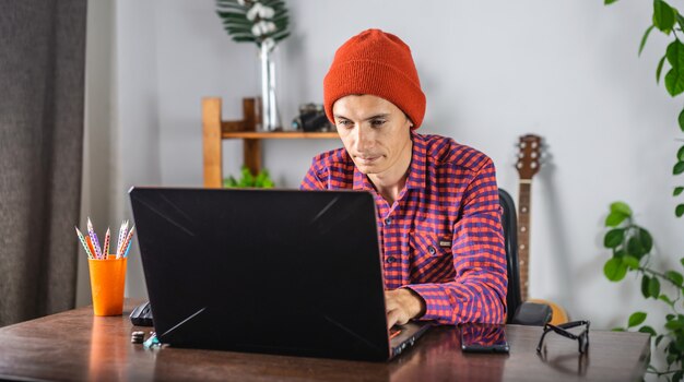 Il giovane moderno in una camicia e un cappello controllati rossi sta lavorando su un computer portatile