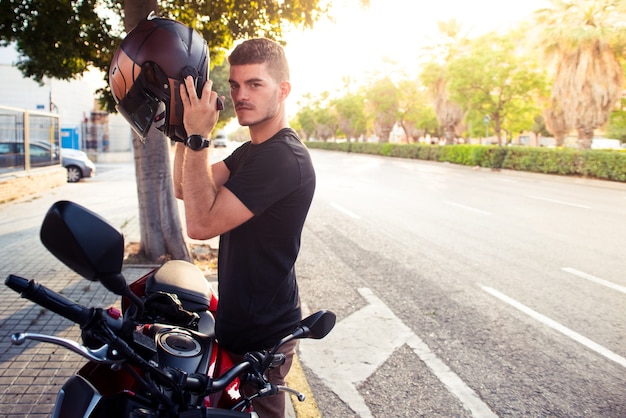 Il giovane motociclista guarda la telecamera mentre indossa il casco per guidare una moto. Concetto di mobilità sicura con moto in città con spazio per il testo.