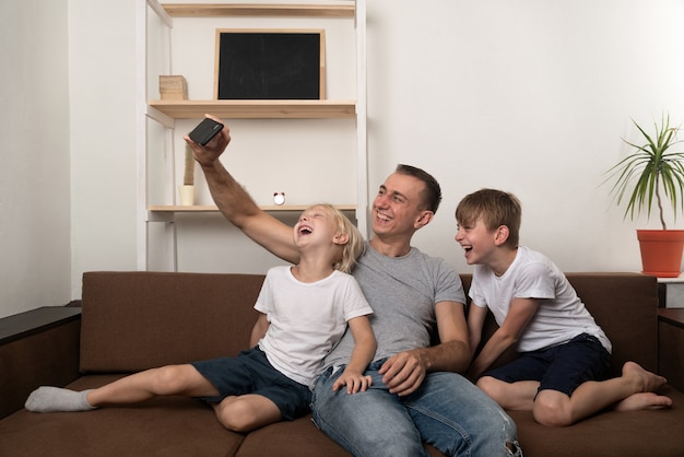 Il giovane padre e due figli si fotografano sullo smartphone sul divano di casa. Tempo divertente