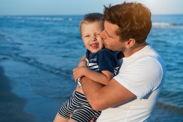 Il giovane papà caucasico con il piccolo figlio cammina la calda giornata estiva lungo la costa di mare. Concetto di vacanze estive in famiglia. Amicizia padre e figlio. Felice infanzia in spiaggia con i genitori. spazio libero per il testo