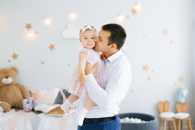 Il giovane papà felice tiene una figlia sorridente tra le braccia e la bacia sulla guancia nella stanza dei bambini