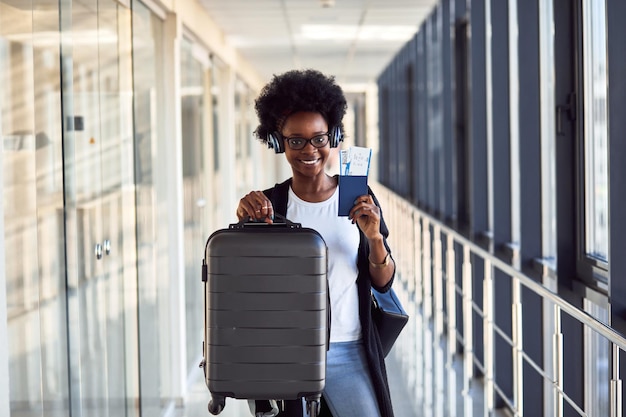 Il giovane passeggero femminile afroamericano in abiti casual e cuffie è in aeroporto con i bagagli.