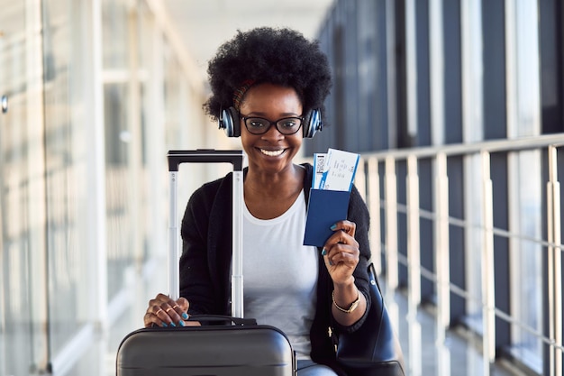 Il giovane passeggero femminile afroamericano in abiti casual e cuffie è in aeroporto con i bagagli.
