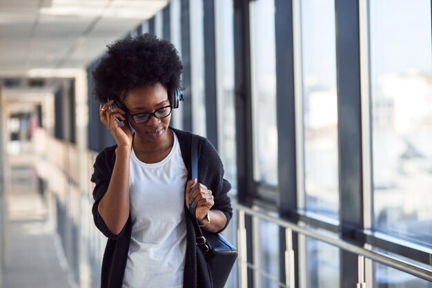 Il giovane passeggero femminile afroamericano in abiti casual è in aeroporto ascoltando musica in cuffia.