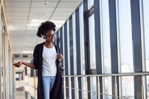 Il giovane passeggero femminile afroamericano in abiti casual è in aeroporto ascoltando musica in cuffia.