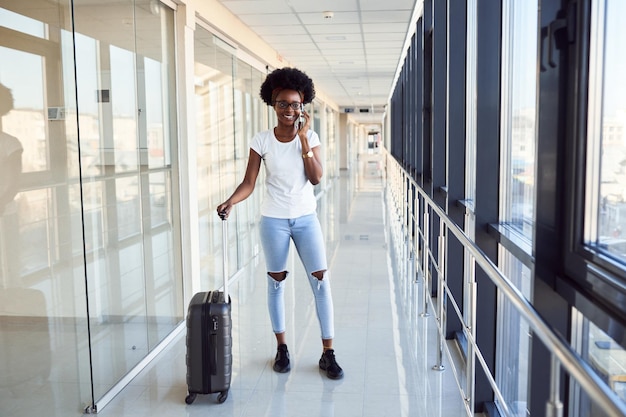 Il giovane passeggero femminile afroamericano in abiti casual è in aeroporto con i bagagli. Usando il telefono.