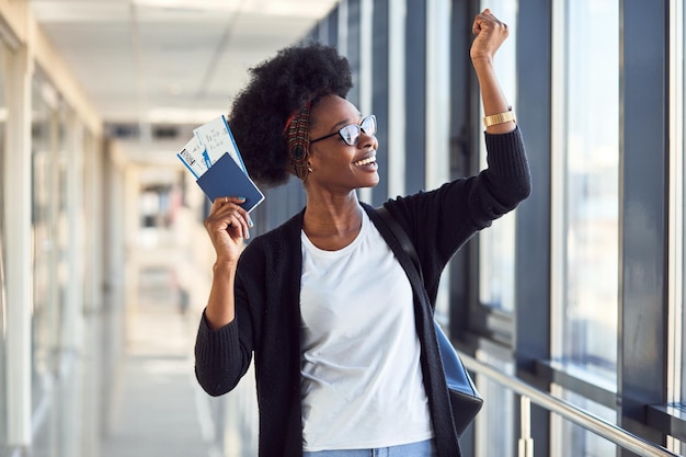 Il giovane passeggero femminile afroamericano in abiti casual è in aeroporto con i biglietti.