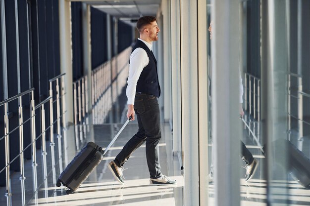 Il giovane passeggero maschio in eleganti abiti formali è nella hall dell'aeroporto che cammina con i bagagli.