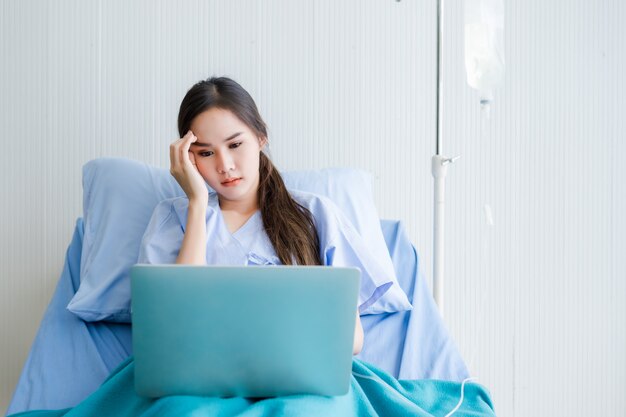 Il giovane paziente femminile asiatico ha mal di testa sul letto e sul computer portatile nell'ospedale della stanza