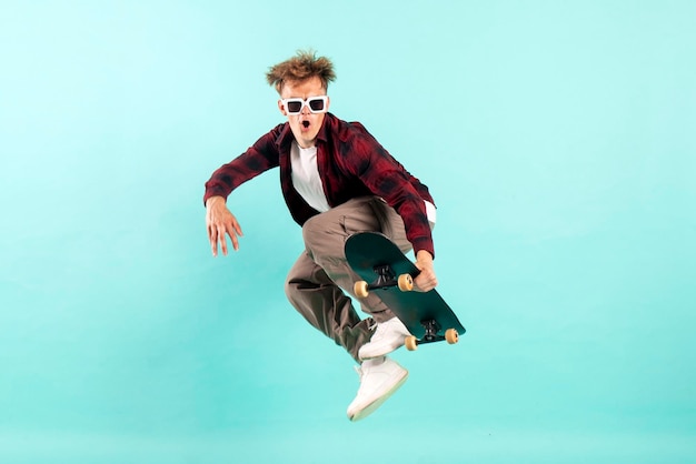 il giovane pazzo cavalca lo skateboard e salta su sfondo blu isolato hipster in occhiali da sole vola