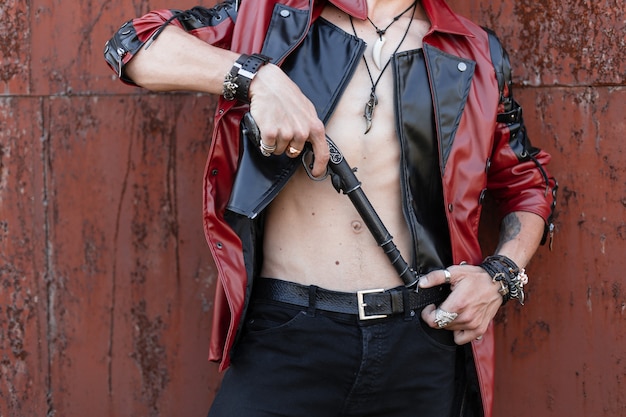 Il giovane pericoloso in un'elegante giacca di pelle in jeans neri con amuleti con bracciali con arma vintage è in piedi vicino a un muro arrugginito all'aperto. Primo piano di un corpo maschile con un torso nudo.
