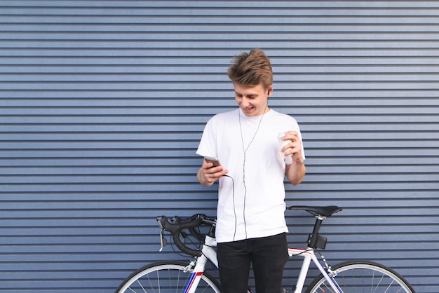 Il giovane positivo sta accanto a una bicicletta contro il muro guarda lo smartphone e sorride