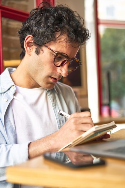 Il giovane prende appunti mentre lavora al computer portatile nell'accogliente caffetteria