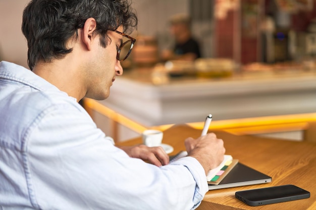 Il giovane prende appunti mentre lavora al computer portatile nell'accogliente caffetteria