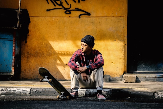 Il giovane ragazzo alla moda con lo skateboard si siede sul marciapiede