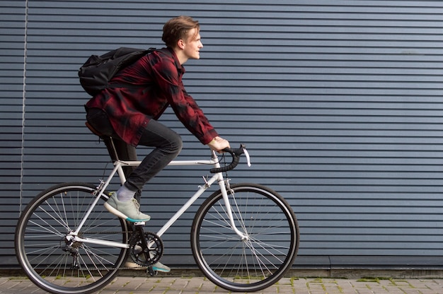 Il giovane ragazzo alla moda guida una bicicletta bianca su una parete a strisce grigia